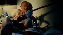 A shirtless man in blue rubber gloves holds a guitar neck in a dimly lit kitchen.