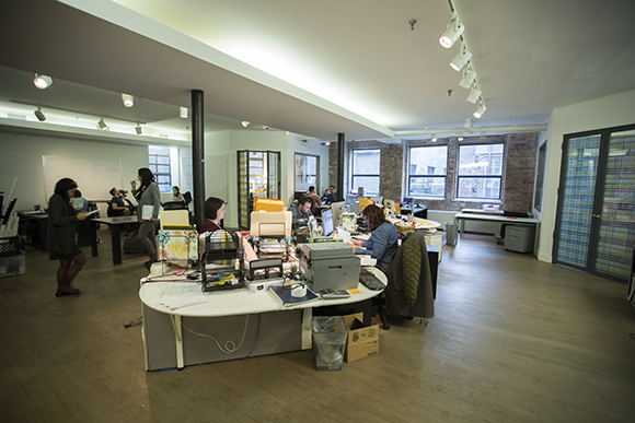 People working at desks in a bright, modern open-plan office with large windows and track lighting.