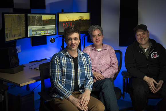 Three men sit in chairs in a studio with computer monitors displaying film editing software.