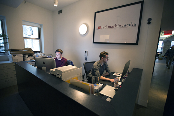Two men sit at desks with computers in an office with a Red Marble Media sign on the wall.