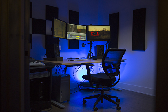 Video editing suite with three monitors, studio monitors, a computer tower, and a black mesh office chair.