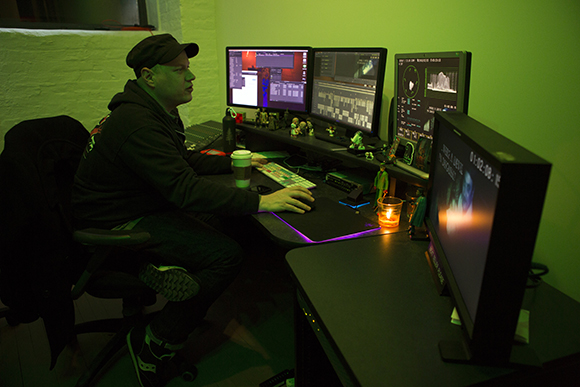 A man in a black hoodie and cap sits at a desk with three monitors displaying data and one monitor in the foreground.