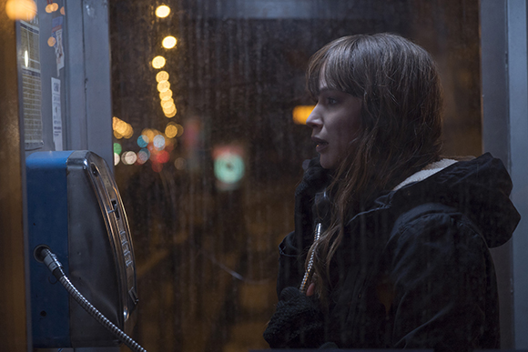 A woman in a dark jacket speaks into a payphone behind a rain-streaked glass window at night.