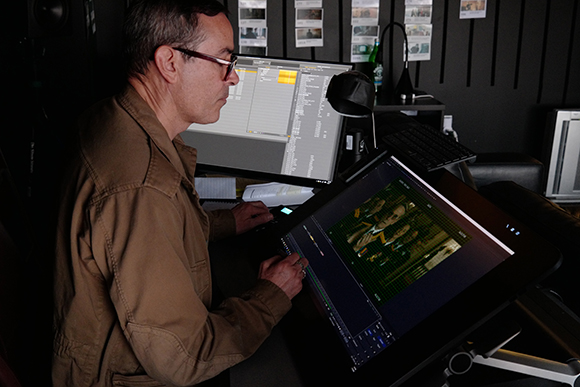 A man in a brown jacket uses a large touchscreen interface and a computer monitor in a dark studio.