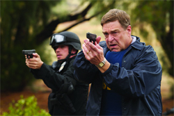 A man in a blue jacket and a man in a black helmet aim handguns toward the camera in a wooded area.