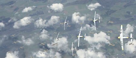 Several white gliders fly in formation over green fields and scattered white clouds.
