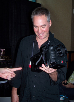 A man in a black collared shirt smiles while holding a black professional camera with a lens attached.