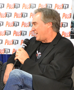 A man with graying hair wearing a black suit jacket over a t-shirt and jeans speaks into a microphone. He is seated in front