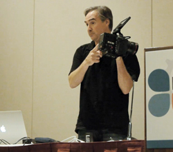 A man in a black shirt holding and looking through a large professional video camera.