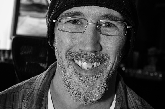 Black and white close-up portrait of a smiling man wearing glasses and a knit beanie.