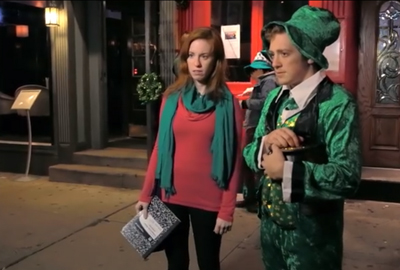 A woman in a red top and green scarf stands beside a man dressed in a green leprechaun outfit.