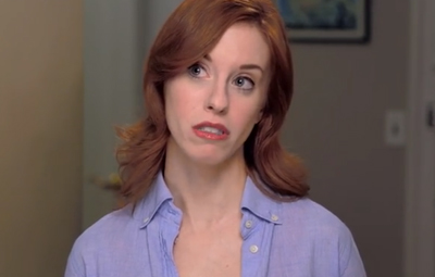 A woman with wavy red hair and a blue button-down shirt looks to the side with a worried expression.