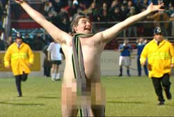 A shirtless man with a striped scarf around his neck celebrates with arms wide on a grass field.