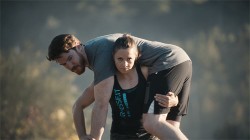 A man in a grey shirt carries a woman in a black tank top on his back outdoors.