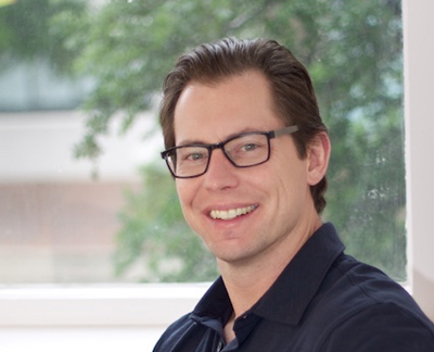 A man with short brown hair and glasses smiling in front of a window with green foliage.