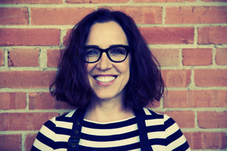 A smiling woman with dark wavy hair and glasses wearing a striped shirt in front of a red brick wall.