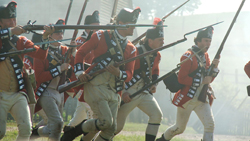 Reenactors in red colonial military uniforms charging forward with muskets and bayonets.