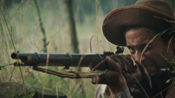 A man in a wide-brimmed hat aims a rifle through tall grass in a wooded area.