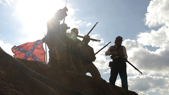 Silhouetted soldiers holding rifles on a grassy hill under a bright sky with a Confederate flag.