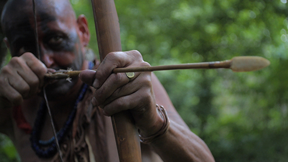 A man with dark face paint aims a wooden bow and arrow in a forest.