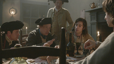 Men in 18th-century attire sit and stand around a wooden table lit by candlelight.
