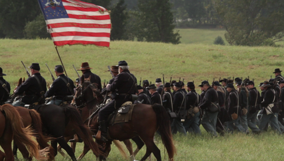 Civil War reenactors in dark uniforms march and ride horses through a grassy field under a large flag.