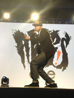 A man in a baseball cap and jacket raps into a microphone on a stage, with a large graphic of a roaring animal behind him.