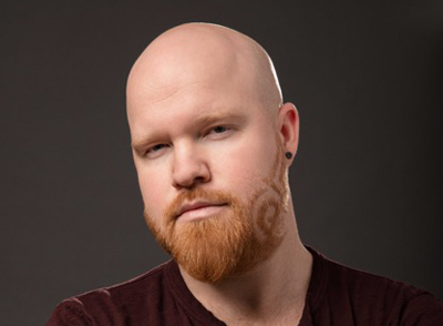 A bald man with a groomed red beard and a small black earring looks toward the camera against a dark background.