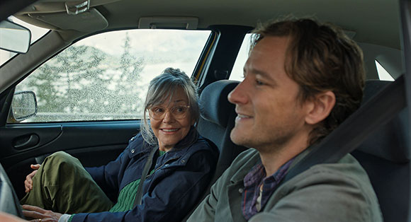 An older woman with grey hair smiles at a man sitting in the front seat of a car.