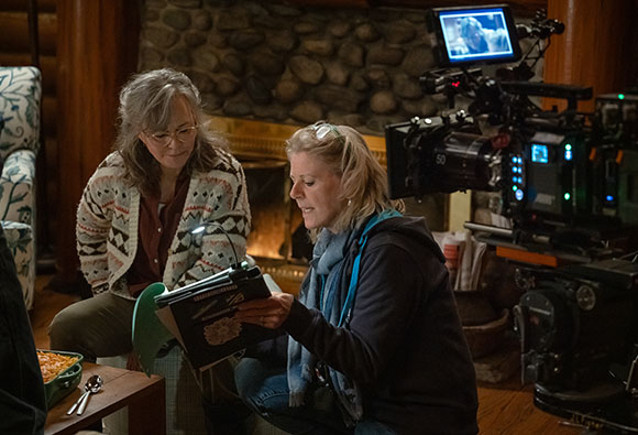 A woman in a patterned cardigan and a woman in a blue scarf review a script near a film camera.