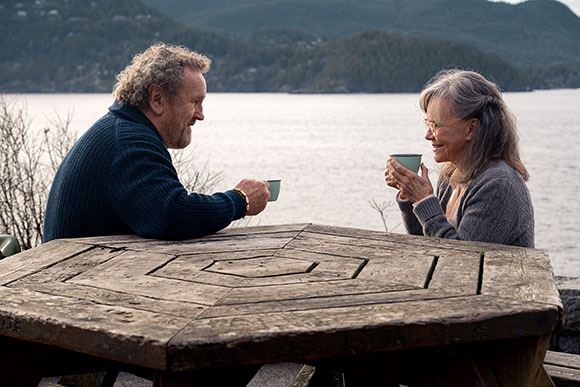 An older man and woman sit at an octagonal wooden table by a lake, smiling and holding small cups.