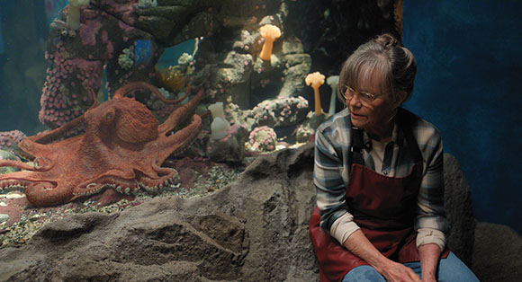 An older woman with glasses and gray hair sits in front of a large aquarium tank. A large red octopus is visible on the left