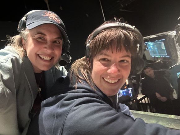 Two women wearing headphones smile for a selfie in a dark studio with production equipment in the background.