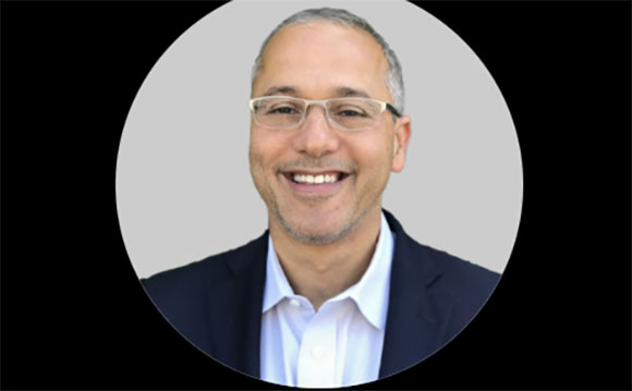 A smiling man with short graying hair and glasses wearing a white shirt and dark blazer.