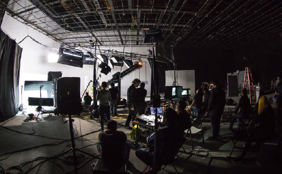 A film crew works on a dark studio set surrounded by professional lighting rigs and cameras.