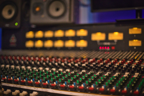 Close-up of a large audio mixing console with numerous colorful knobs and sliders in a studio setting.