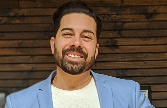 A smiling man with dark hair and a beard wearing a light blue blazer and white shirt.