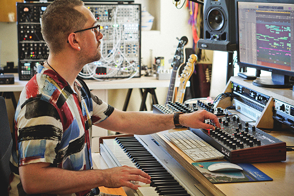 A man in a patterned shirt sits at a desk using a MIDI controller and keyboard in a music studio.