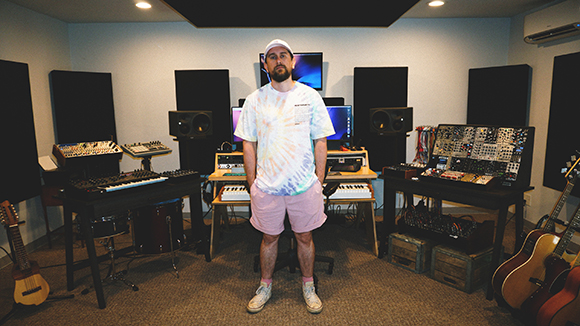 A man in a white t-shirt and pink shorts stands in a music studio filled with synthesizers and instruments.