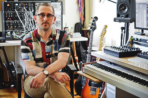 A man in a patterned shirt sits in a music studio filled with synthesizers, guitars, and audio gear.