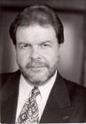 Black and white portrait of a man with dark hair and a beard wearing a suit and patterned tie.