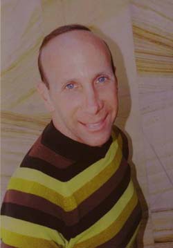 A smiling man with blue eyes and a receding hairline wearing a brown and yellow striped turtleneck.