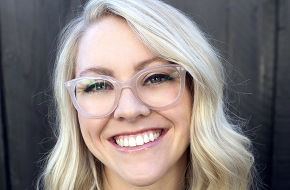 A smiling woman with wavy blonde hair and clear-framed glasses.