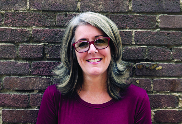 A smiling woman with shoulder-length brown hair and glasses wearing a maroon shirt against a brick wall.
