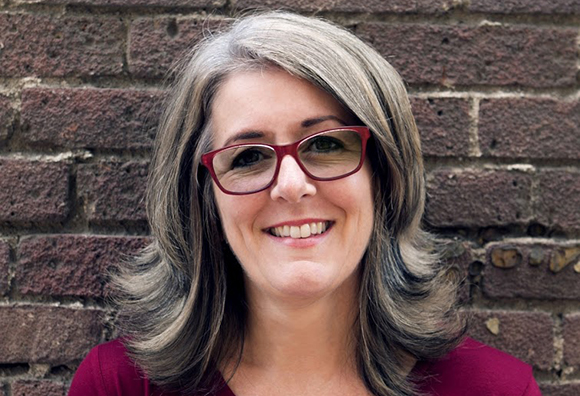 A smiling woman with shoulder-length grey hair and red glasses posing in front of a brick wall.