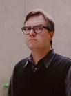 A man with glasses and short brown hair wearing a dark sweater vest over a collared shirt.