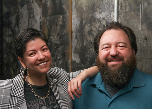 A smiling woman in a plaid blazer leans her arm on a smiling man with a thick brown beard.