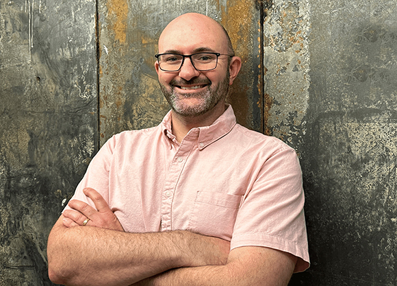 A smiling man with glasses and a beard wearing a pink short-sleeved shirt, standing with arms crossed.