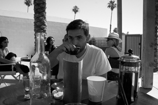 A man sits at an outdoor table drinking from a small cup, surrounded by people and palm trees.