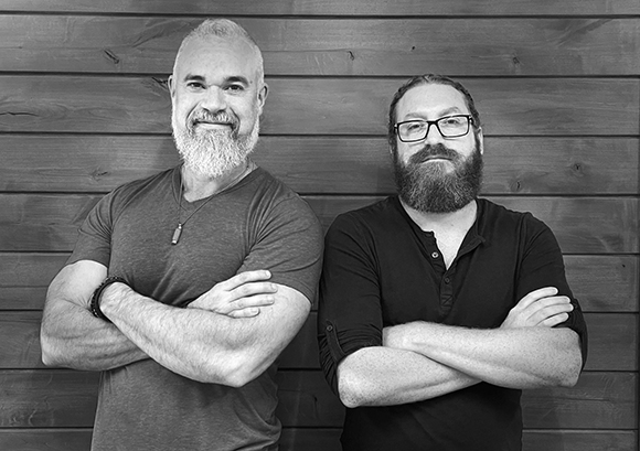 Black and white portrait of two bearded men standing side by side with arms crossed against a wood wall.